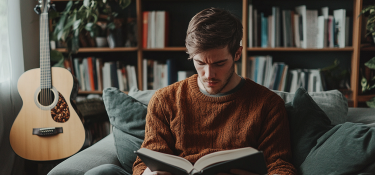 A creative man reading a book on a couch with a guitar leaning against a shelf behind him